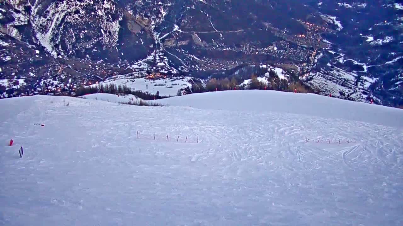 A snow-covered slope shows ski tracks leading down to a valley with a village at twilight.