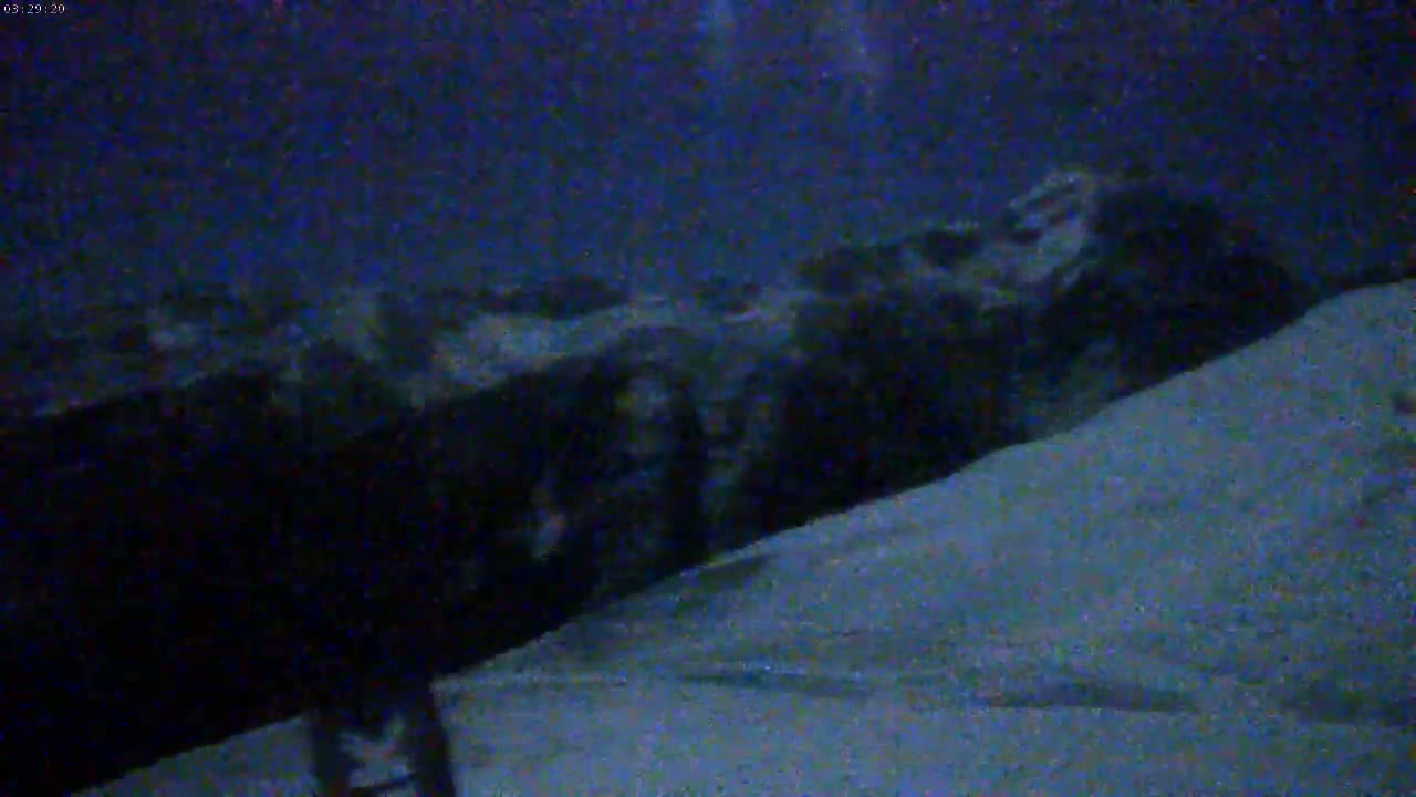 A dark, low-resolution image shows a large, dark object, possibly a vehicle or structure, partially submerged in or adjacent to a snow-covered area at night.