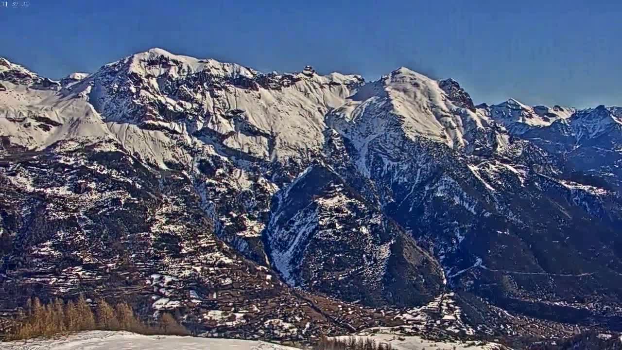 A snow-covered mountain range stretches across a clear blue sky, with a valley below showing some sparse vegetation and a small settlement.