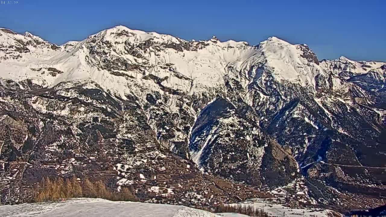 A snow-covered mountain range is seen on a clear, sunny day, with a valley below containing a small town partially visible among dark trees.