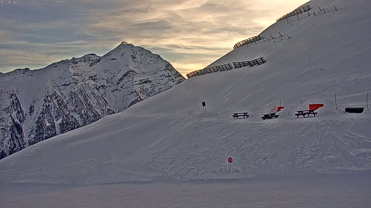 A snow-covered mountain slope with picnic tables and avalanche barriers at sunset.