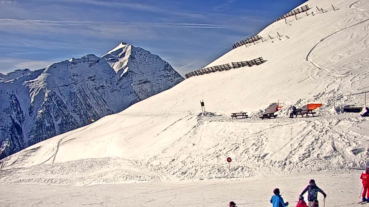 A snowy mountain slope with a few skiers and picnic tables, under a bright, sunny sky.