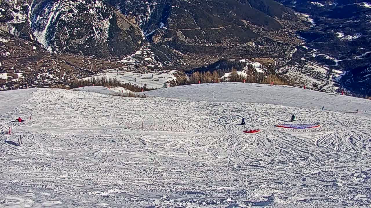 A snowy mountain slope with several people and a paraglider, overlooking a valley on a sunny day.