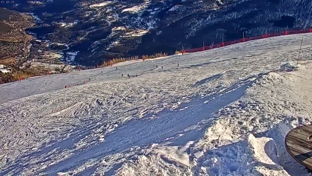 A snowy ski slope with several skiers visible on a sunny day, overlooking a mountainous valley.
