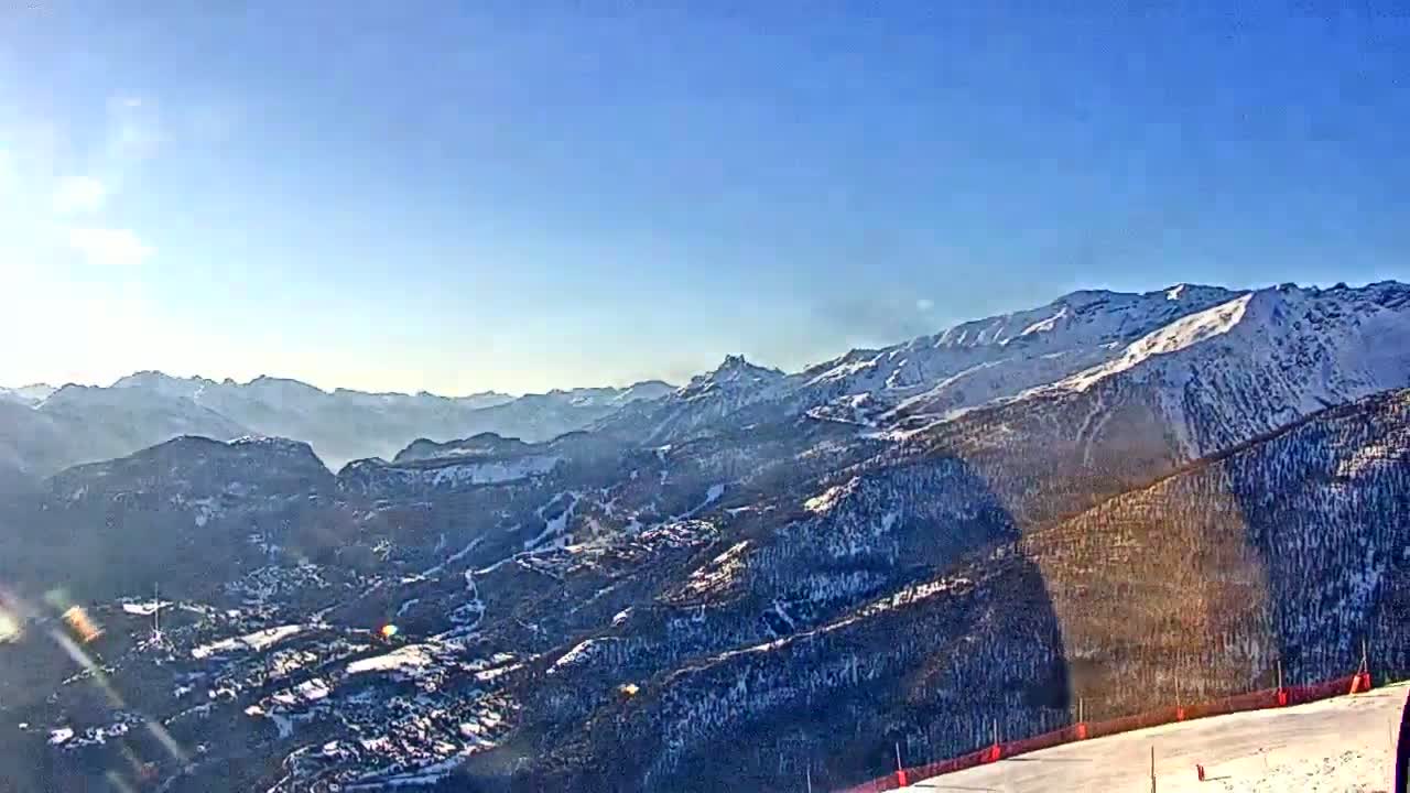 A sunny, snow-covered mountain range stretches into the distance, with a ski slope visible in the foreground.