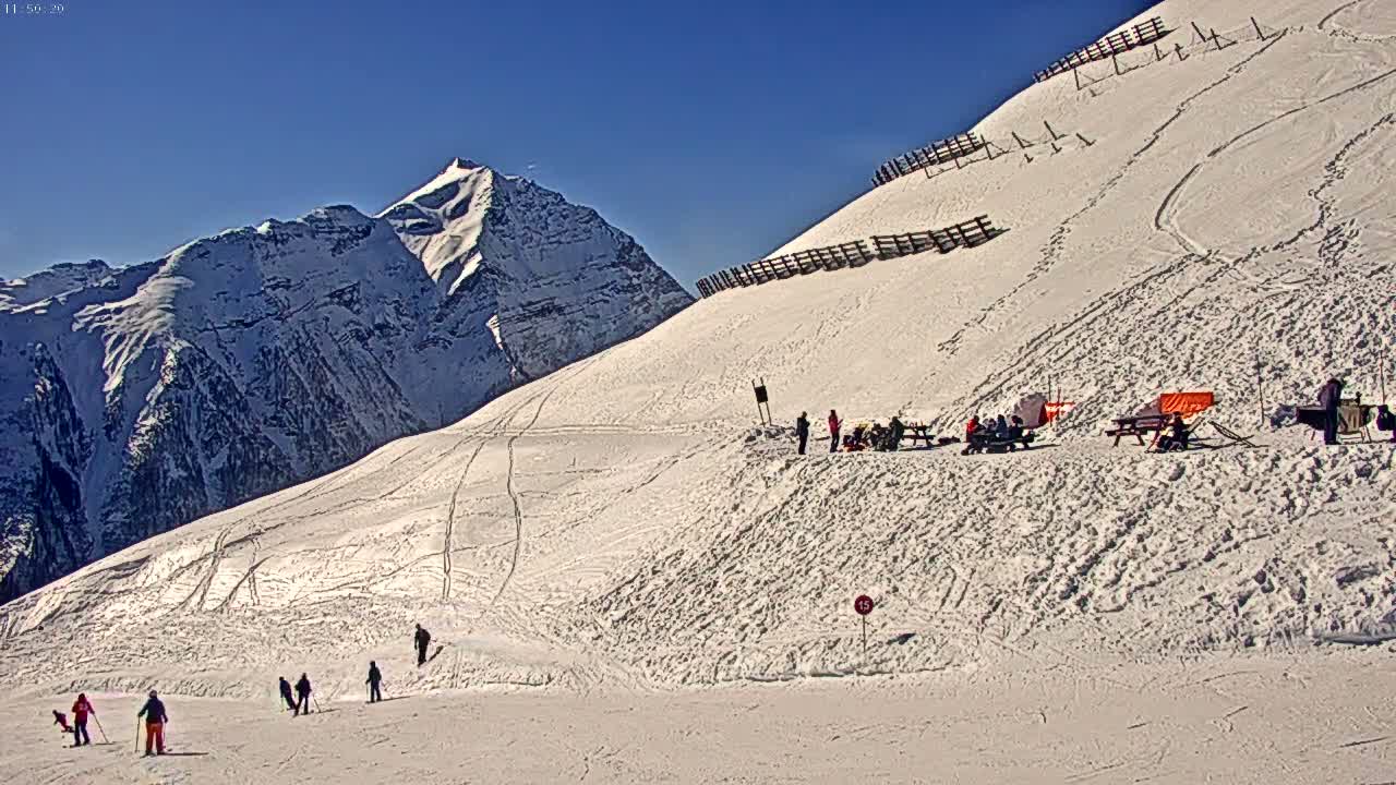 A sunny, snow-covered mountain slope shows skiers and snowboarders enjoying the winter day, with a mountain range in the background.