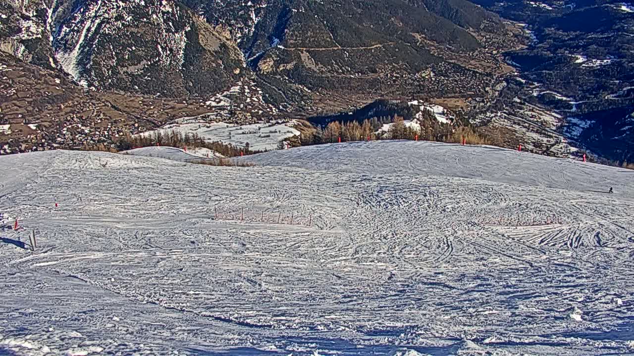 A snow-covered ski slope with many ski tracks is situated on a mountain overlooking a valley on a sunny day.