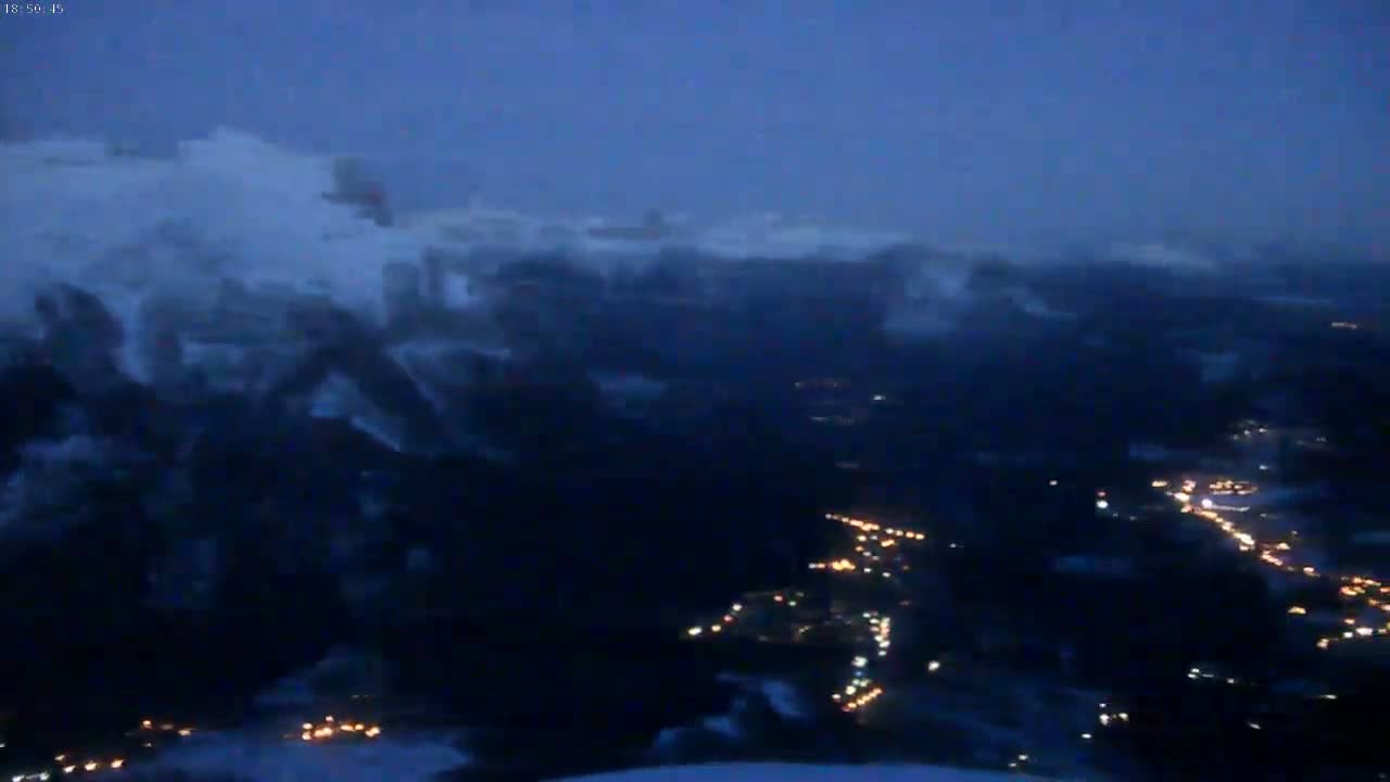 A nighttime, snowy mountain range is visible with a town's lights scattered below.