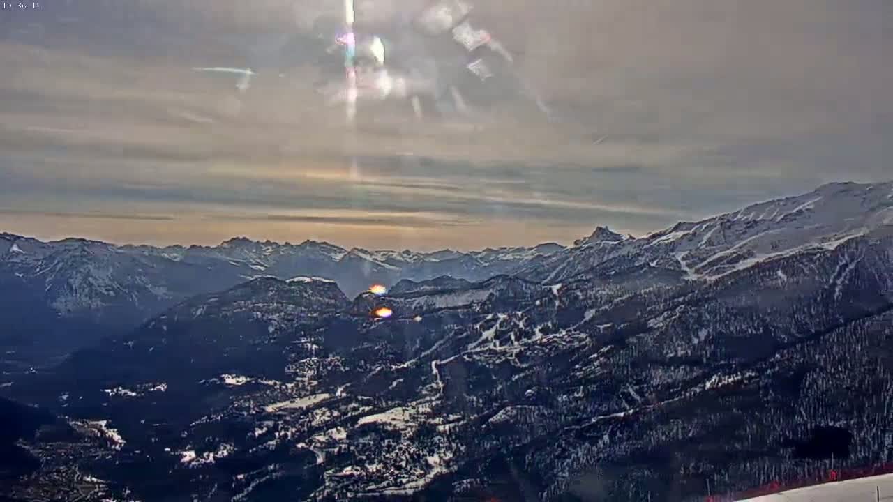 A sunlit, expansive view of snow-covered mountains and valleys under a partly cloudy sky.