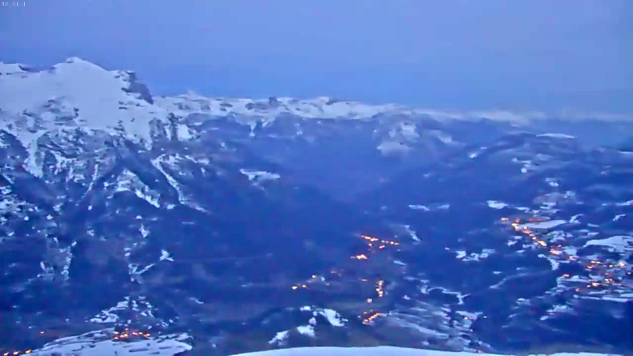 A snow-covered mountain range at dusk overlooks a valley with scattered lights below.