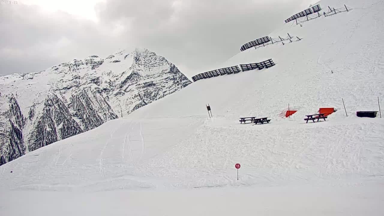 A snow-covered mountain slope with several picnic tables and avalanche barriers under an overcast sky.