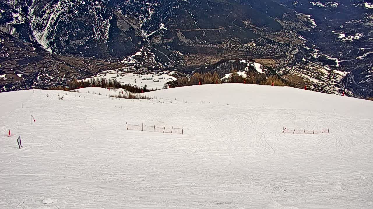 A snow-covered ski slope overlooks a valley with dark green mountains under a bright, sunny sky.