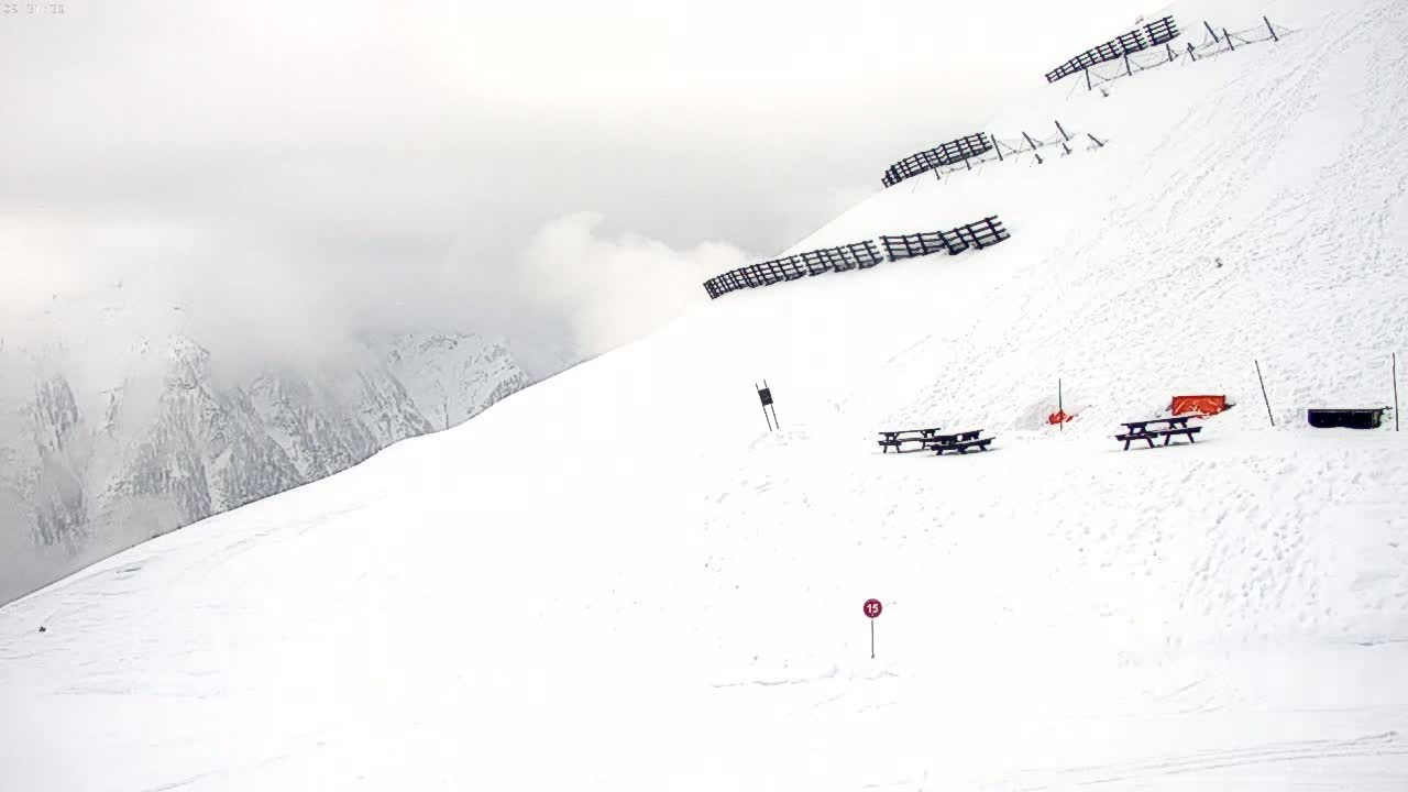 A snow-covered mountain slope with picnic tables and avalanche fencing is visible under an overcast sky.