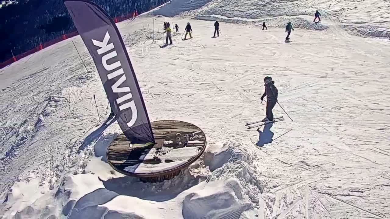 A sunny day on a snow-covered ski slope shows several skiers and snowboarders, with one skier near a round wooden structure next to a tall vertical banner.