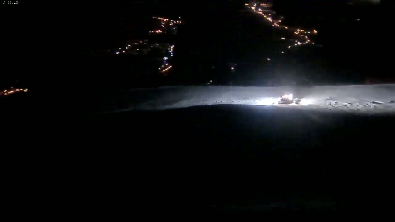 A snowcat is grooming a snow-covered slope at night, with a town's lights visible in the distance.