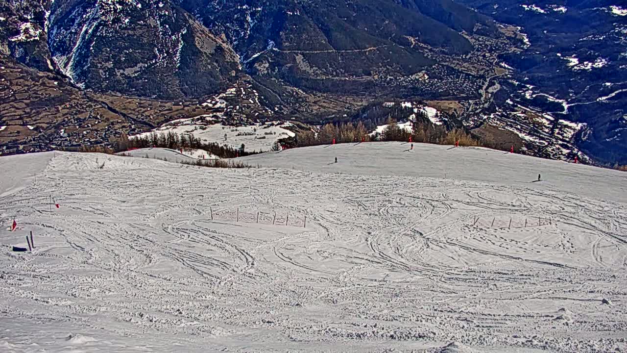 A snow-covered ski slope with numerous ski tracks is situated on a mountainside overlooking a valley on a sunny day.