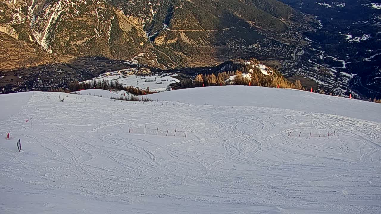A snow-covered ski slope with numerous ski tracks shows a valley and distant mountains in the background under a sunny sky.