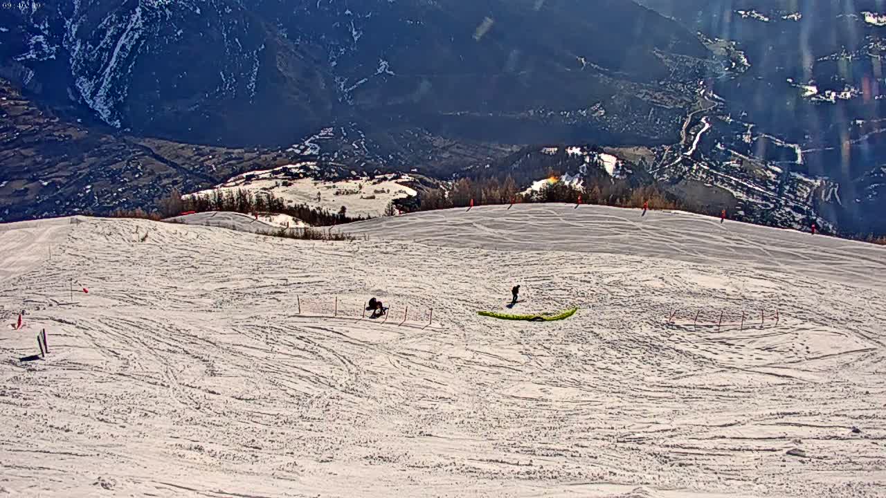 A snowy ski slope with a few skiers and snowboarders, overlooking a valley on a sunny day.
