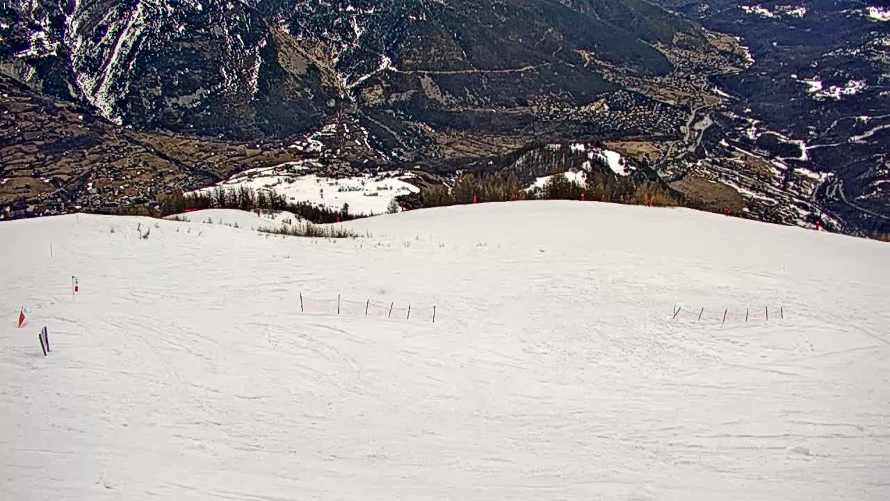 A snow-covered ski slope overlooks a valley with brown, sparsely vegetated hills and mountains in the distance under a clear sky.