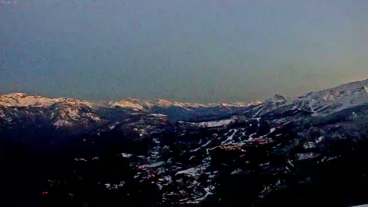 A snow-covered mountain range is visible at dusk, with a valley and town nestled below.