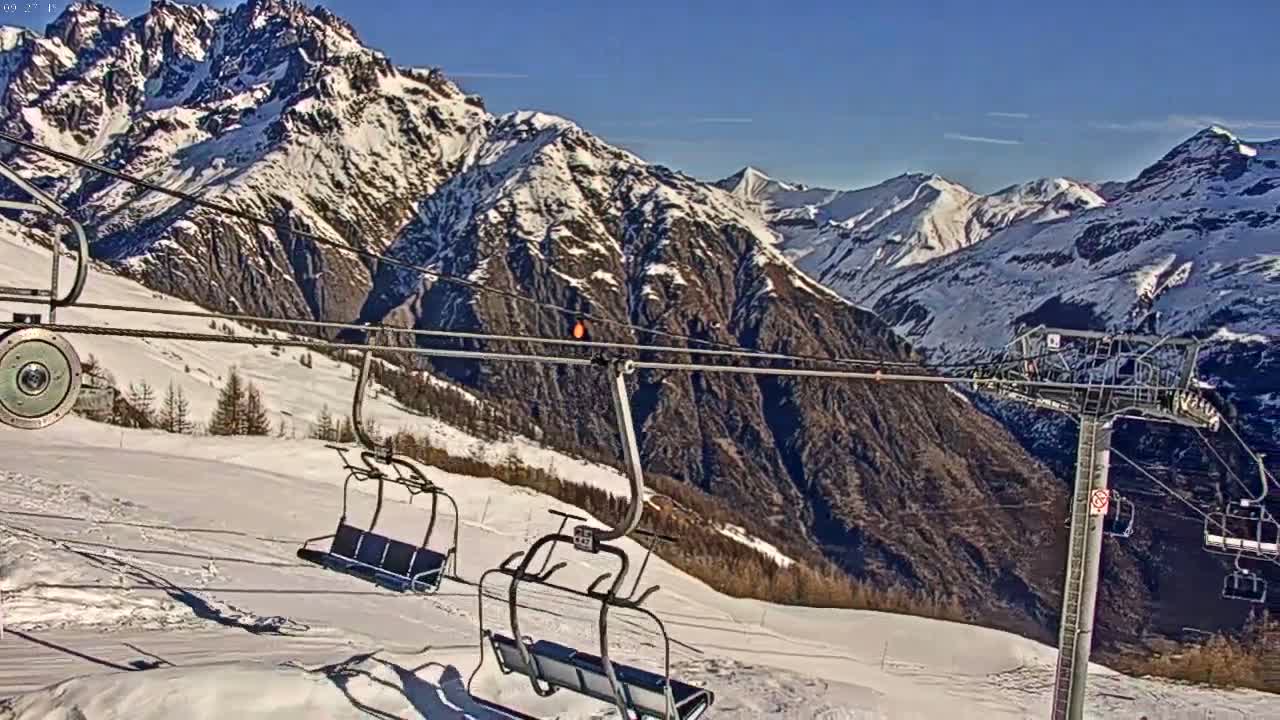 A ski lift with empty chairs overlooks a snow-covered mountain range on a sunny day.