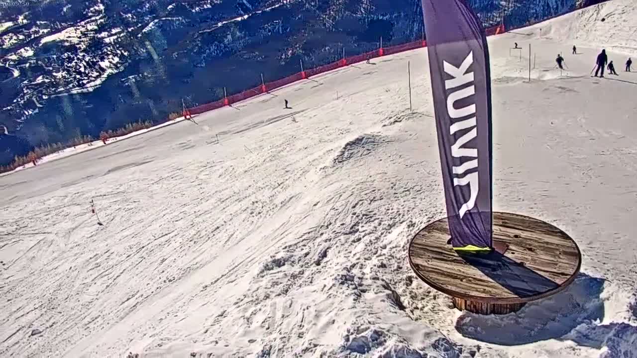 A snowy ski slope with several skiers visible, a tall banner standing on a circular wooden base, and mountains in the background under a sunny sky.