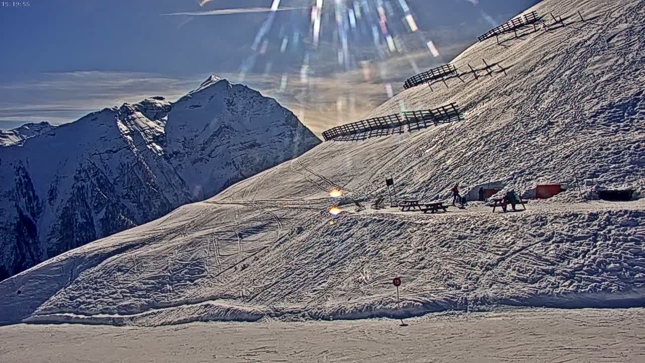 A snowy mountain slope with a few people and picnic tables is bathed in bright sunlight against a clear blue sky.