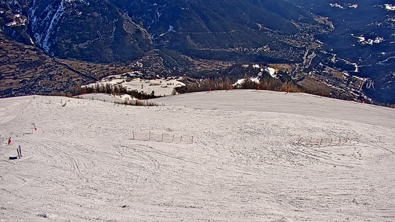 A snowy ski slope with sparse markings overlooks a valley of dark green hills and a village under a bright, sunny sky.