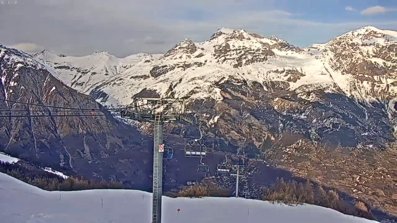 A ski lift stands on a snowy mountainside overlooking a valley and snow-capped mountains under a partly cloudy sky.