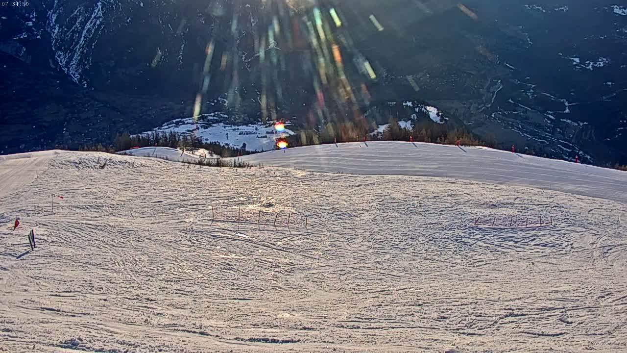 A snowy ski slope with several skiers visible, situated on a mountainside with a valley and sun shining brightly in the background.