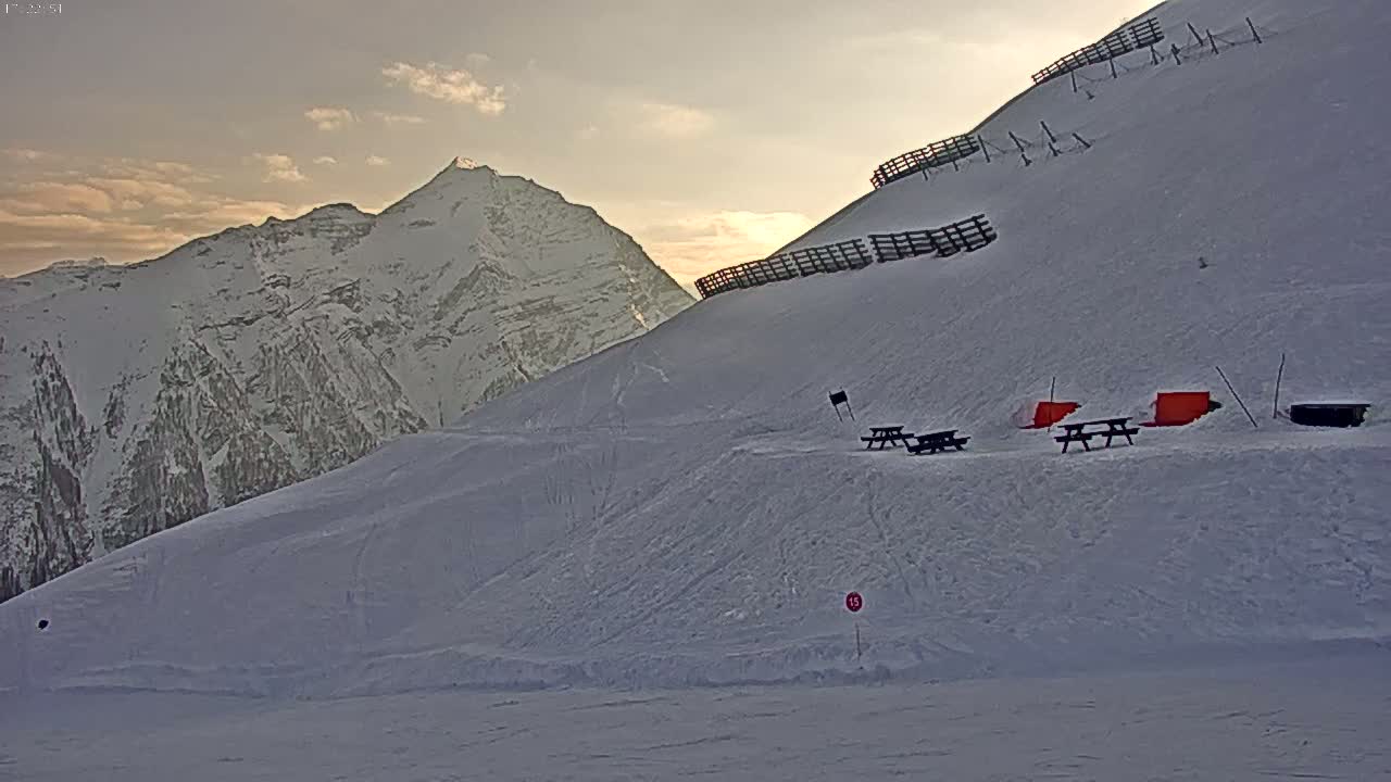 A snow-covered mountain slope with picnic tables, under a partly cloudy sunset sky, features a snow-capped mountain range in the background.