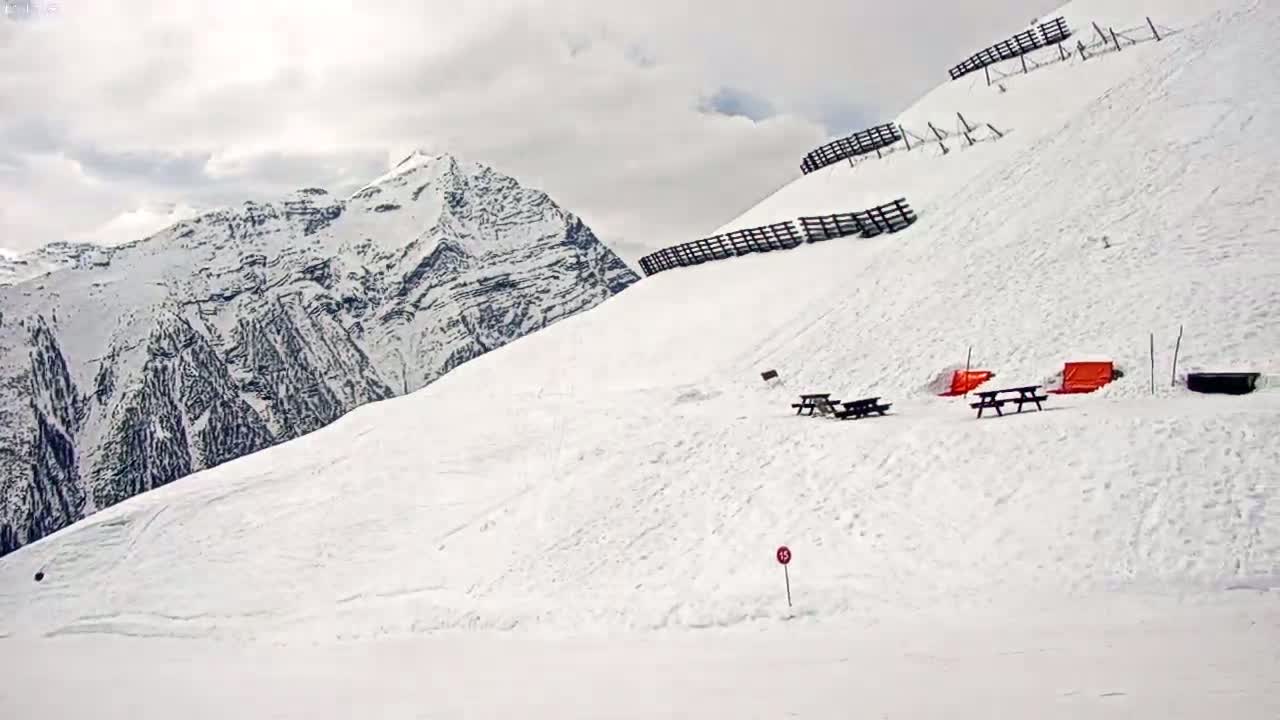 A snow-covered mountain slope with picnic tables, avalanche barriers, and orange structures, under a cloudy sky.