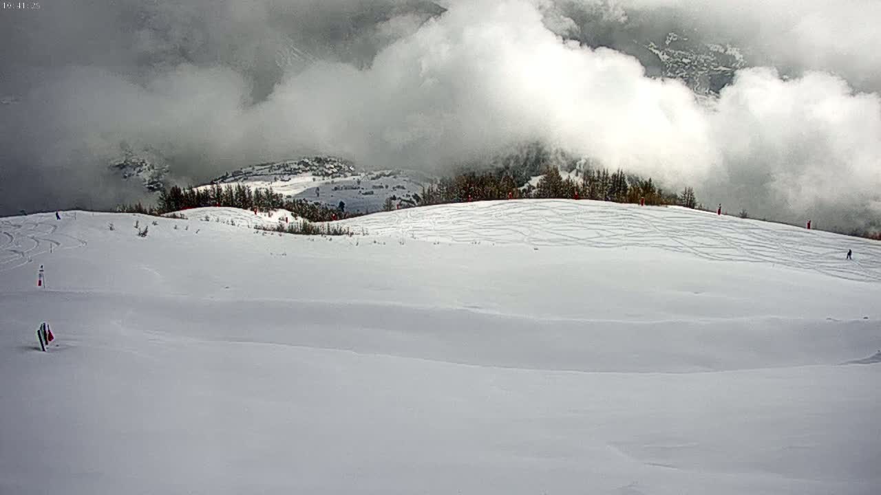 A snowy mountain slope with several skiers visible, partially obscured by low-hanging clouds.