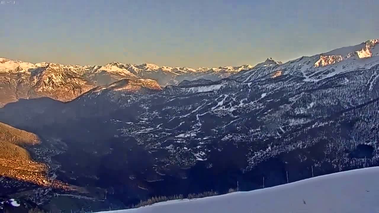 A snow-covered mountain range stretches into the distance under a clear, pale blue sky, with the lower slopes showing patches of dark trees and a valley below.