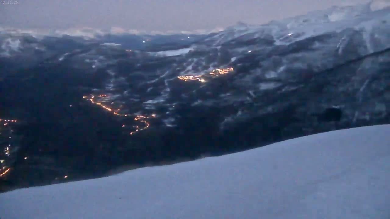 A snow-covered mountain slope overlooks a valley with a town illuminated at night, under a dusky sky.