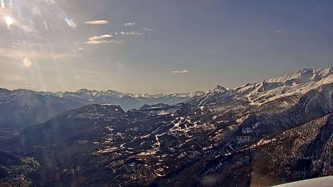 A sunlit view of a snow-capped mountain range and a valley below, with a partly cloudy sky.