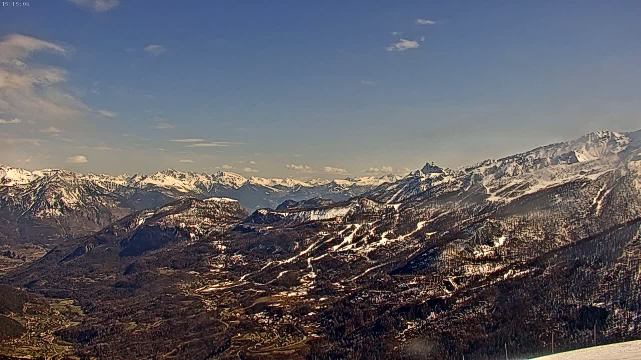 A panoramic view of snow-capped mountains and a valley under a mostly clear, bright sky.
