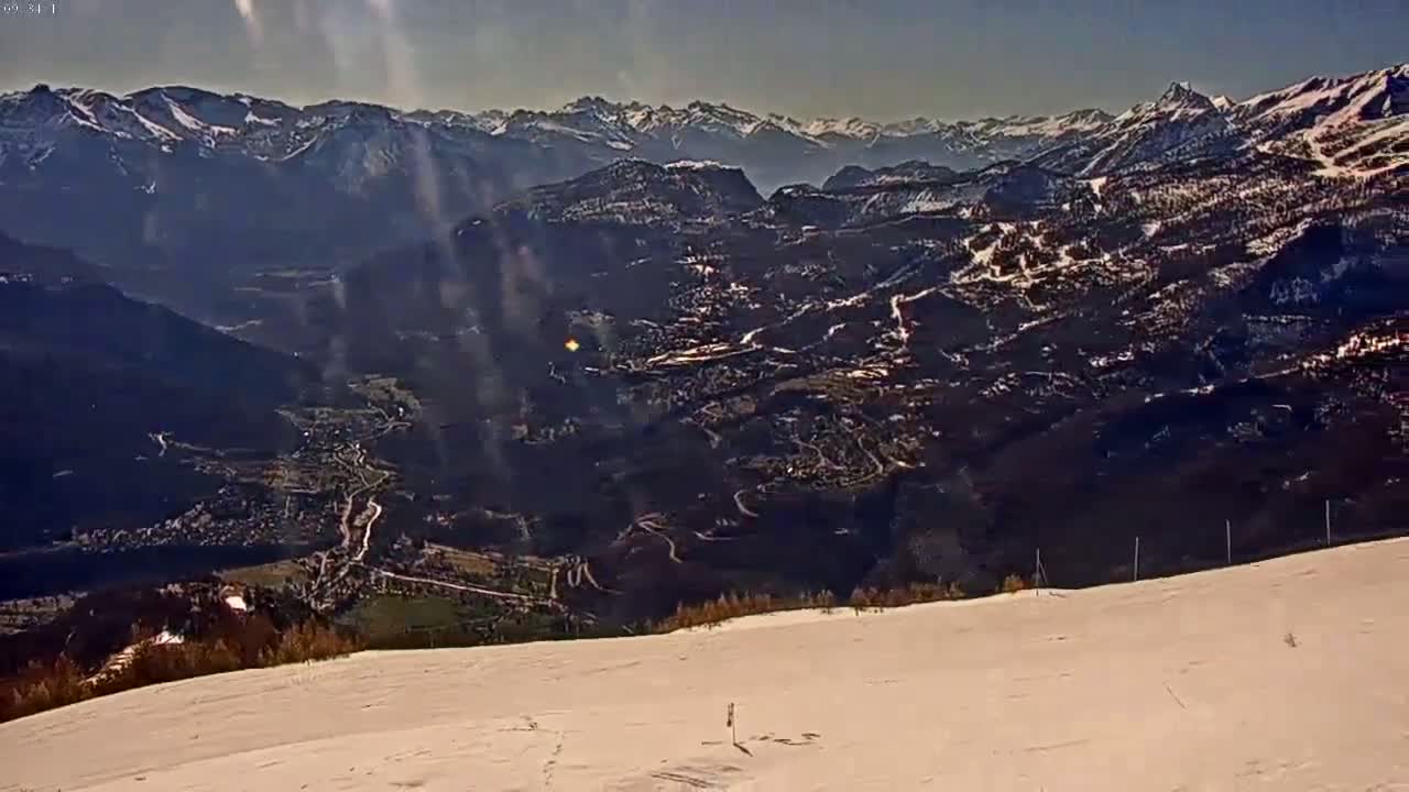 A snow-covered mountaintop overlooks a valley with a town nestled among the dark, snow-dusted hills and mountains under a bright, sunny sky.