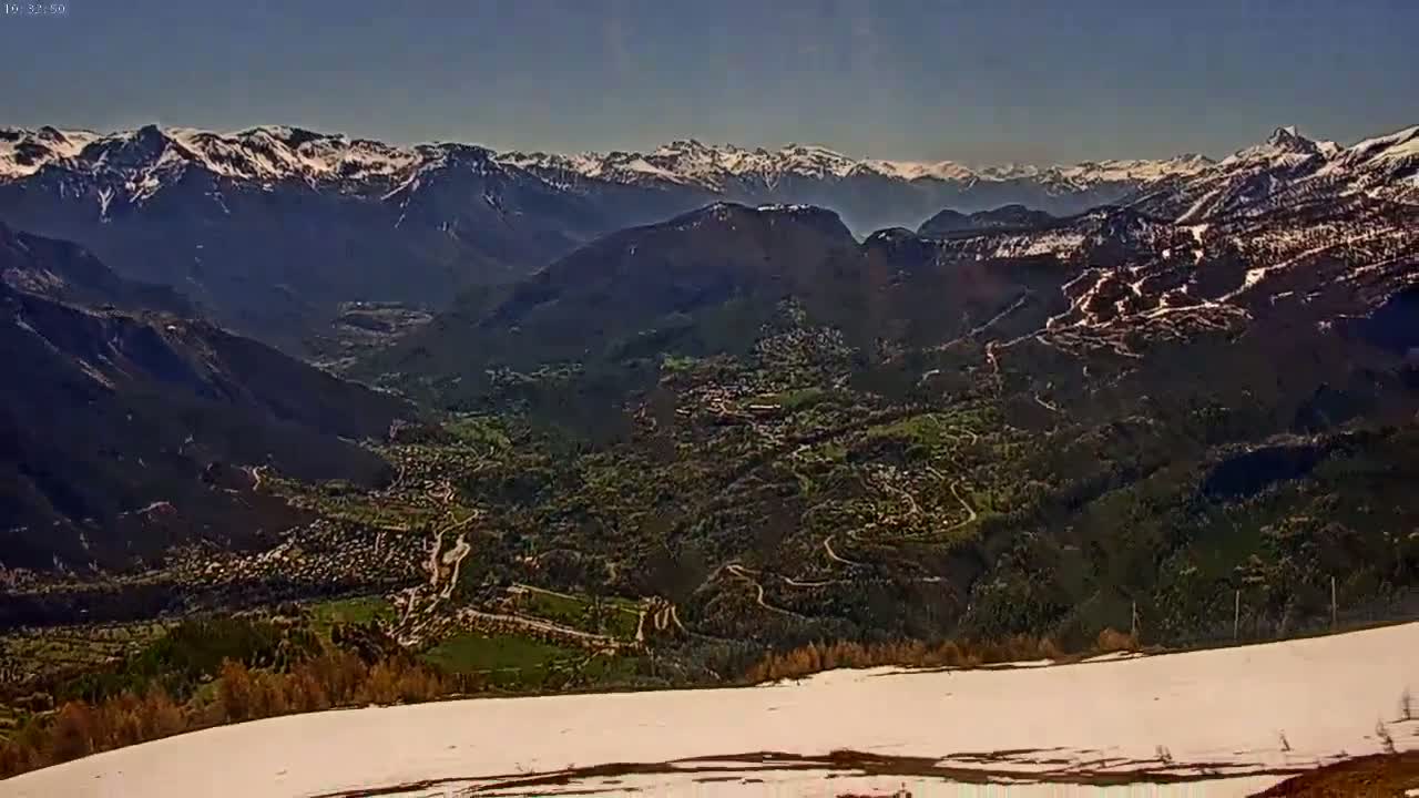 A mountain valley with a town nestled among green hills and winding roads, viewed from a snowy vantage point under a clear blue sky.