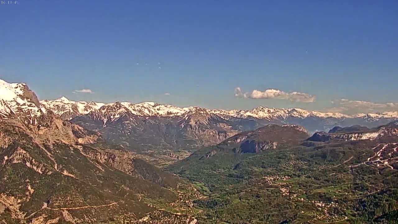 A sunny day reveals a vast mountain range, with snow-capped peaks in the distance and a valley of green and brown hills in the foreground.