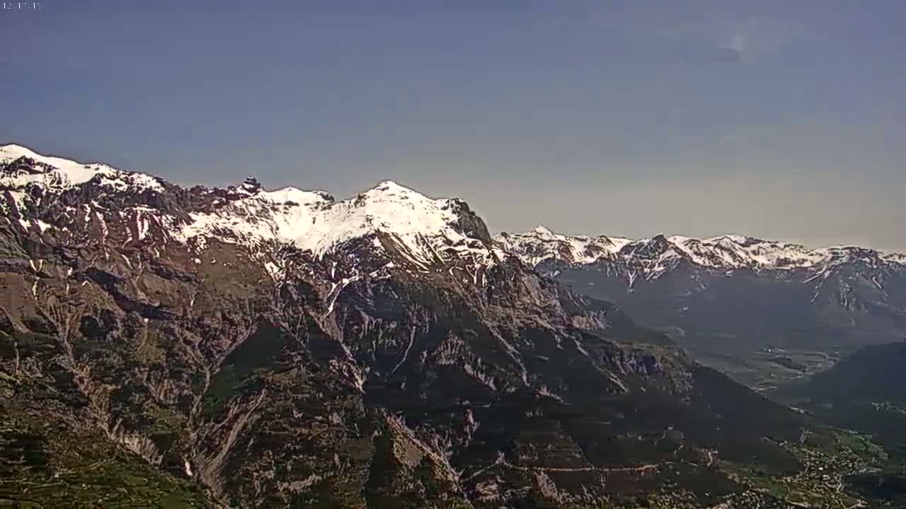 A snow-capped mountain range stretches across a clear, sunny sky, overlooking a valley below.