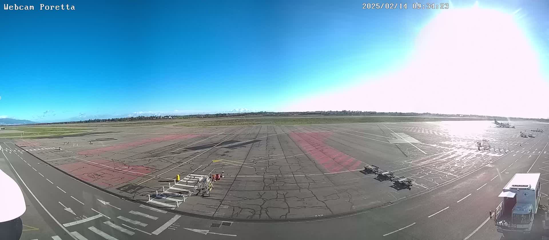 A sunny day at an airport shows a tarmac with several vehicles and ground equipment, a plane parked in the distance, and a clear blue sky.