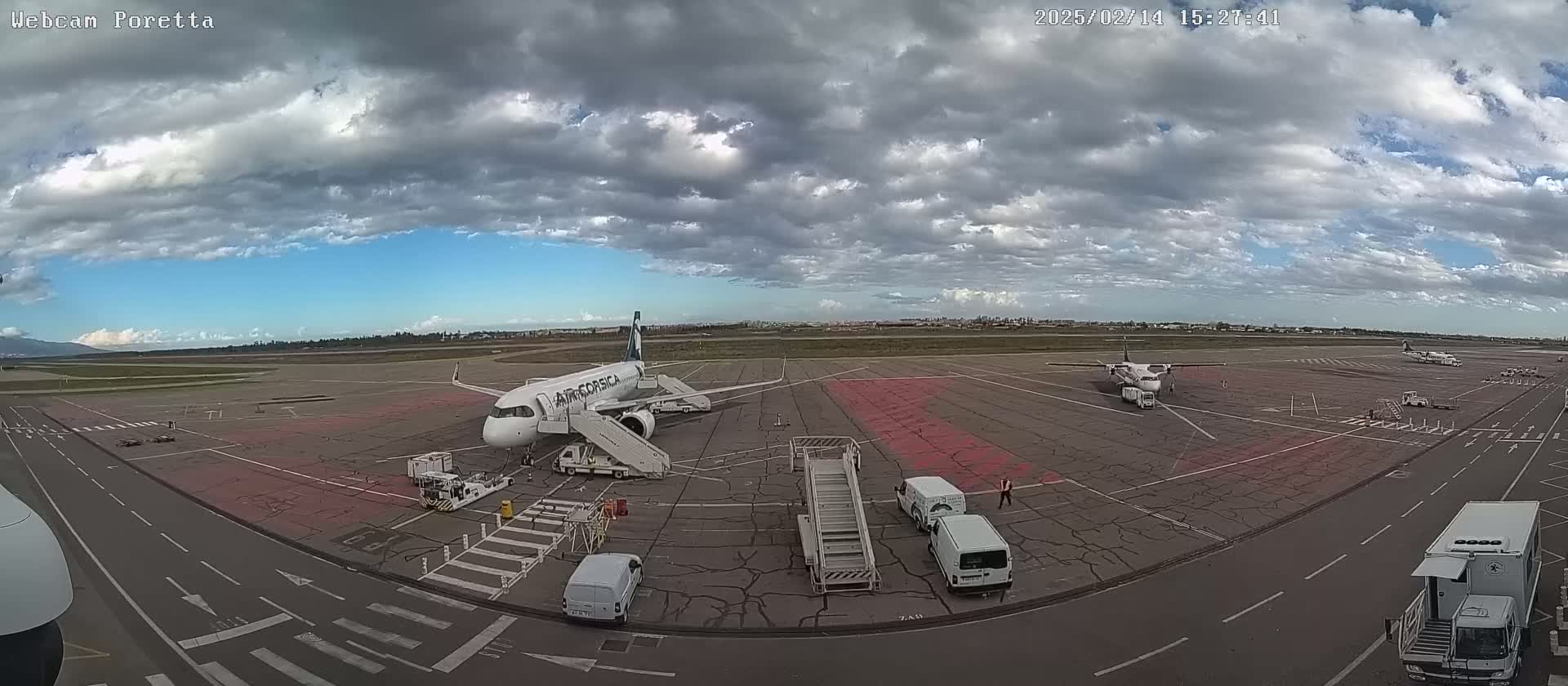 An airport tarmac under a partly cloudy sky shows two airplanes, one with a jet bridge, surrounded by ground support vehicles.