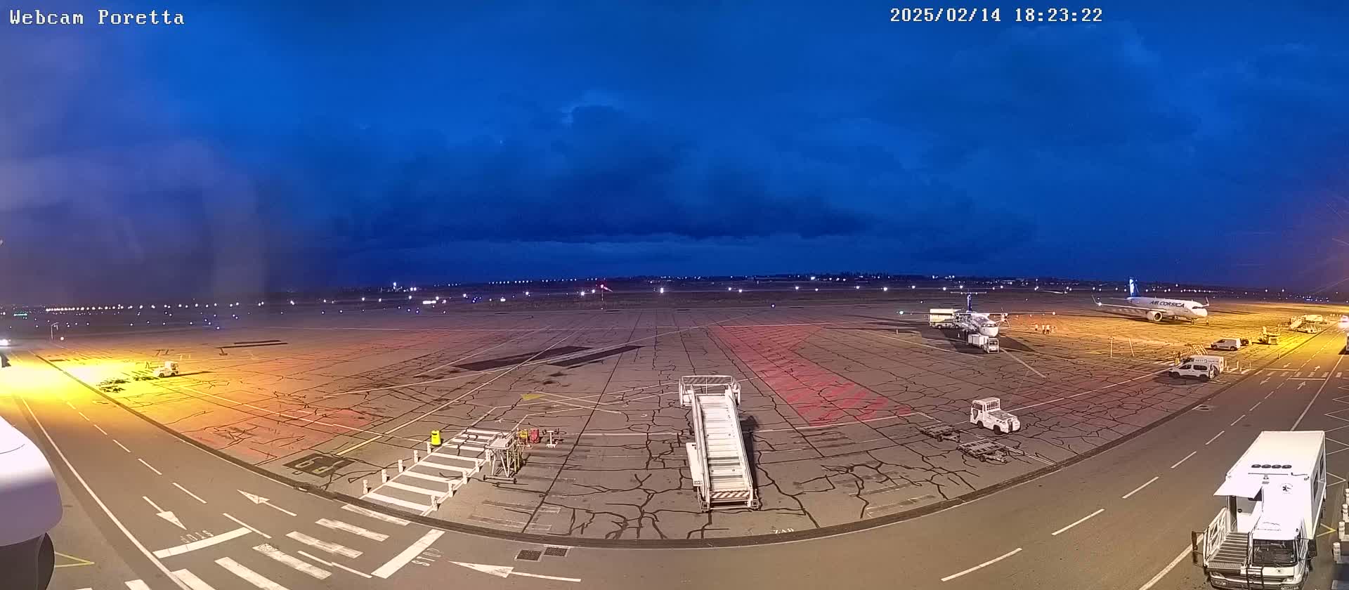 A nighttime view of an airport tarmac with several airplanes, ground support vehicles, and jet bridges under a partly cloudy sky.