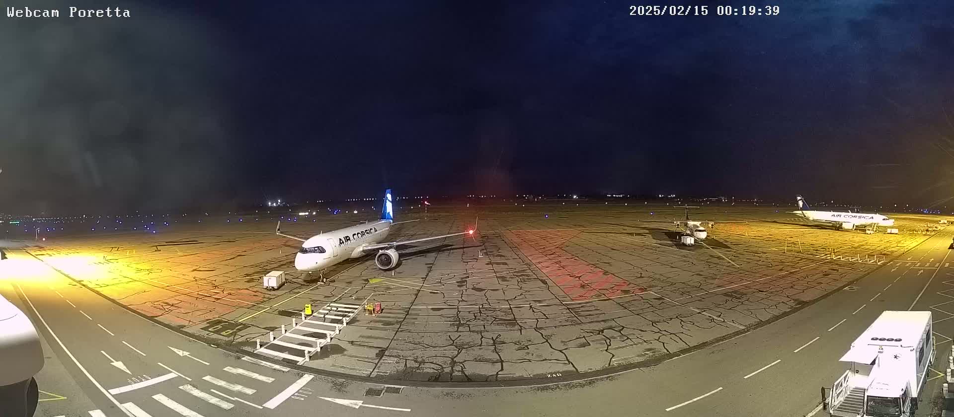At night, a tarmac shows two airplanes parked, one larger than the other, with airport service vehicles present under a dark, mostly clear sky.