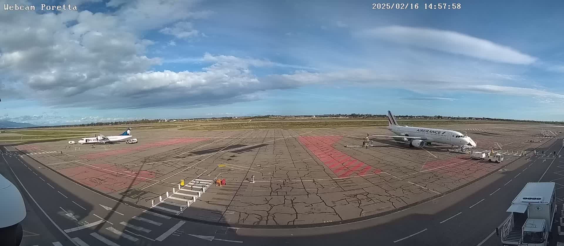 A partly cloudy day at an airport shows two airplanes on the tarmac, one being serviced by ground crew.