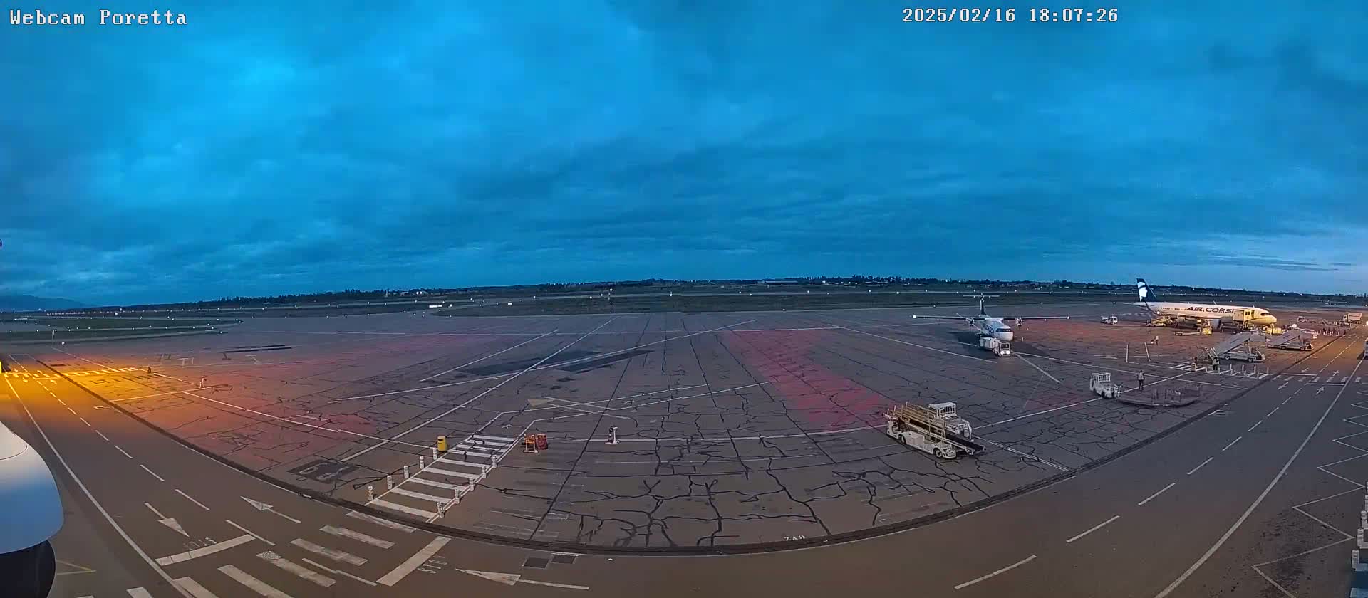 An airport tarmac at twilight shows two airplanes parked at gates with ground support equipment present under a mostly clear, dark blue sky.