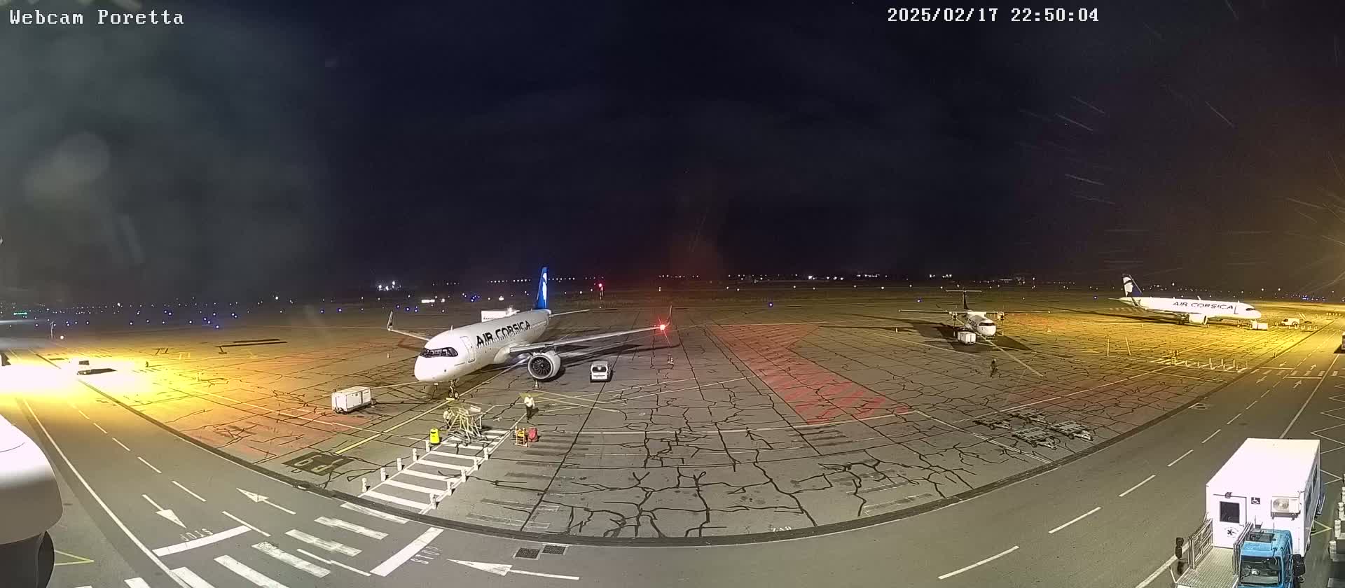 At night, an airport tarmac shows two airplanes parked, one larger than the other, with ground crew present under a dark, clear sky.
