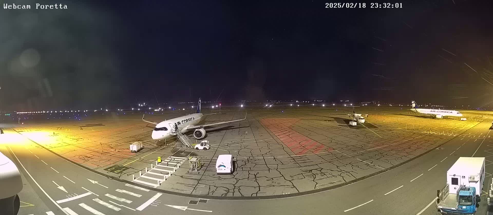 At night, a tarmac at an airport shows two airplanes parked, one with a stairway attached,  surrounded by ground support vehicles and illuminated by runway lights.