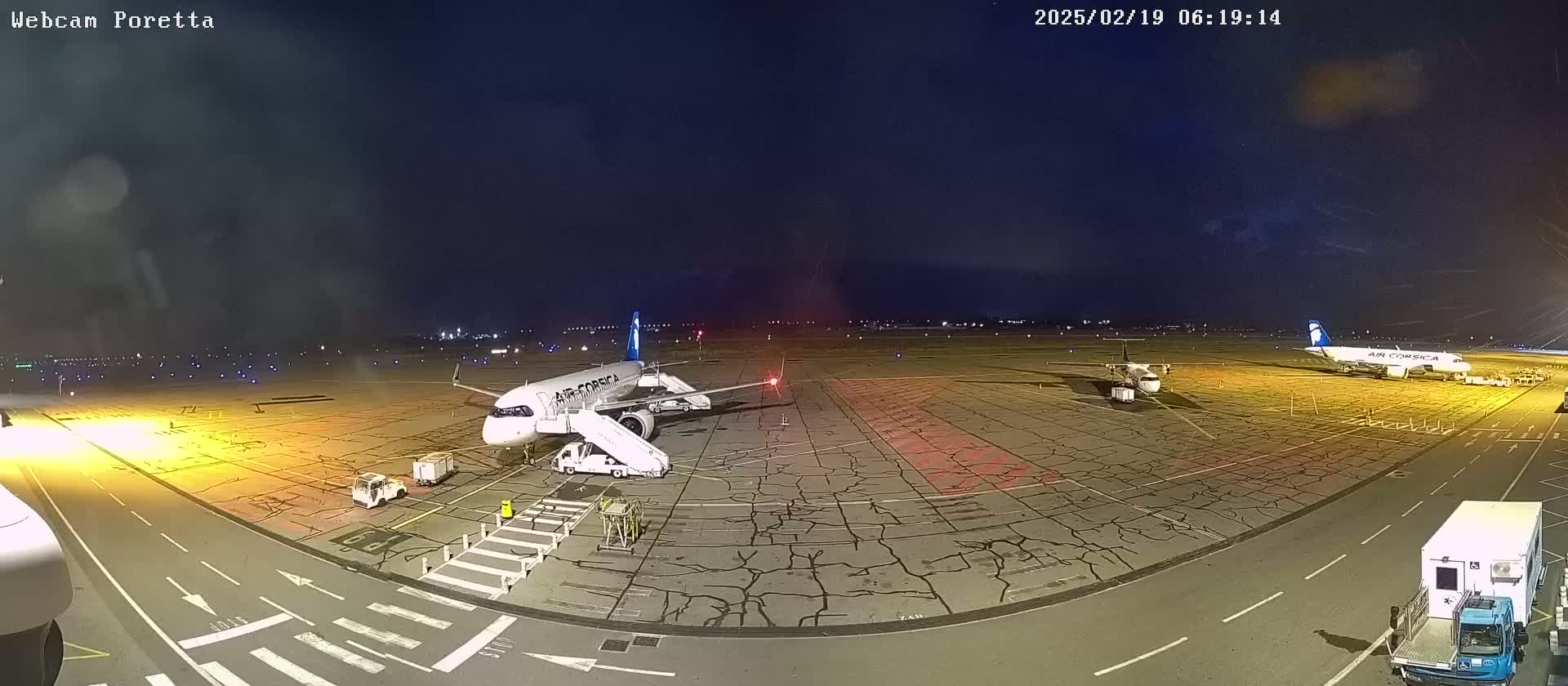 An airport tarmac at night, showing two airplanes parked with ground support equipment, under a dark, slightly hazy sky.